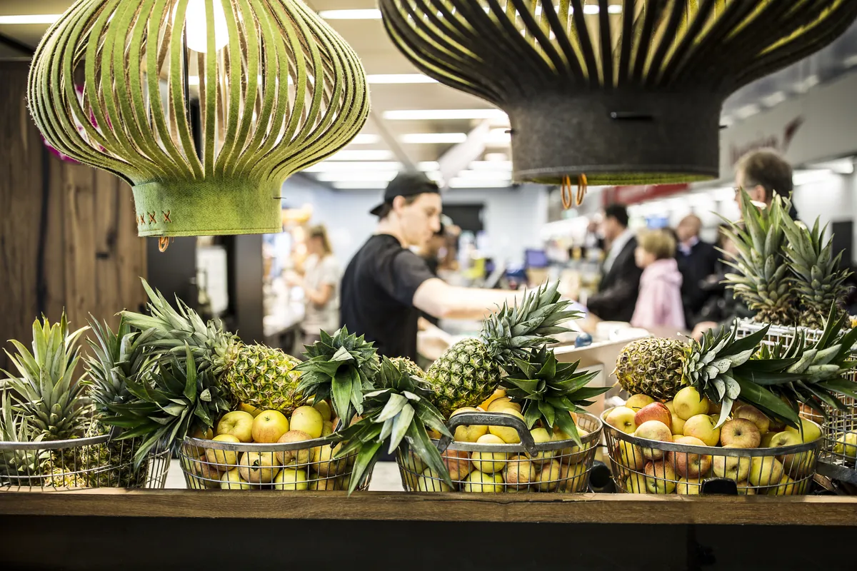 Pineapples and apples in wire baskets under felt lamps, a Juicer at the counter behind them at Vienna Airport