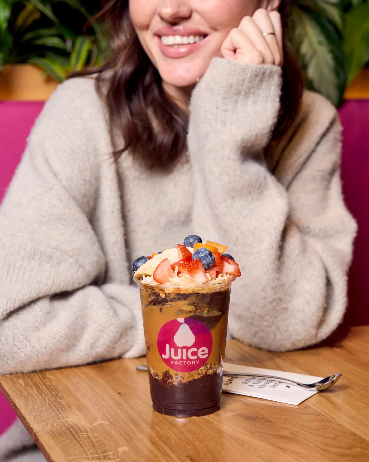 Lächelnde Frau am Tisch mit einem Açaí-Becher der Juice Factory, getoppt mit Beeren und Granola
