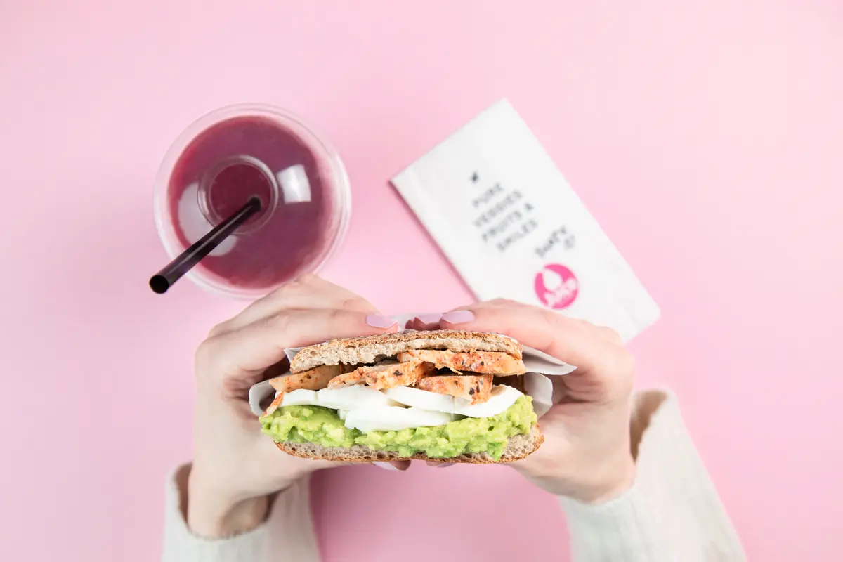 Hands holding a toasted chicken and avocado sandwich next to a berry juice