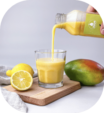 Eine Hand gießt gelben Saft aus einer Flasche in ein Glas auf einem Holzbrett, neben dem Glas stehen auf hellem Hintergrund eine halbierte Zitrone und eine ganze Mango.