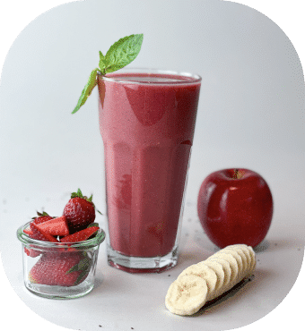 Ein großes Glas Erdbeer-Smoothie, garniert mit einem Minzblatt, daneben ein kleines Glas Erdbeeren, ein roter Apfel und Bananenscheiben auf einem Brett, alles auf einer hellen Oberfläche.
