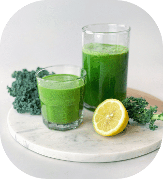 Zwei Gläser mit grünem Smoothie stehen auf einem runden Marmortablett, umgeben von frischen Grünkohlblättern und einer halbierten Zitrone. Die Szene ist hell und klar und betont die gesunden, frischen Zutaten.