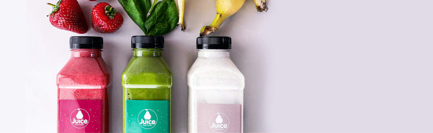 Drei Flaschen mit bunten Saft-Smoothies – rosa, grün und weiß – stehen nebeneinander. Darüber befinden sich Erdbeeren, Spinatblätter und eine Banane auf hellem Hintergrund.