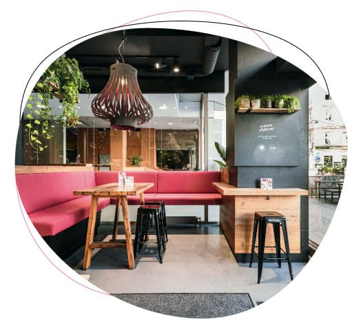 Modern cafe interior with wooden tables, black stools, a pink cushioned bench, hanging plants, and a unique pendant light. Large windows let in natural light, creating a cozy and inviting atmosphere.
