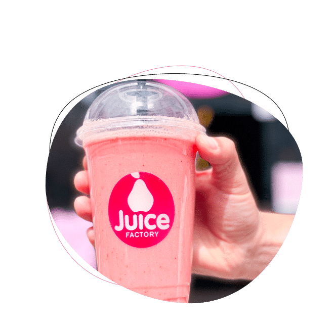 A hand holding a pink smoothie in a clear plastic cup with a dome lid, featuring a Juice Factory logo on the front of the cup.