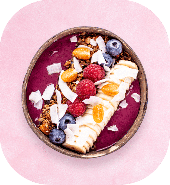 Eine Smoothie-Bowl mit Bananenscheiben, Blaubeeren, Himbeeren, Gelbbeeren, Granola und Kokosflocken, serviert in einer runden Schüssel auf rosa Hintergrund.