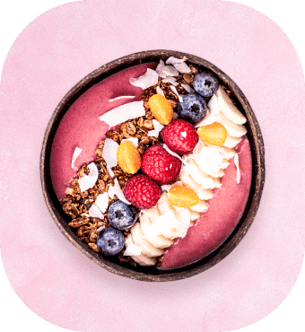 Eine Smoothie-Bowl mit Granola, Bananenscheiben, Himbeeren, Blaubeeren, Gelbbeeren und Kokosflocken, serviert in einer runden Schüssel auf einem rosa Untergrund.