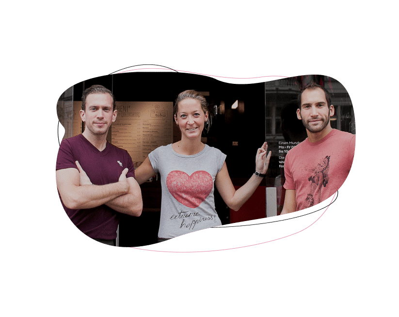 Three people stand smiling in front of a storefront. The woman in the middle wears a light t-shirt with a red heart, and the two men on either side wear maroon t-shirts. All appear happy and relaxed.