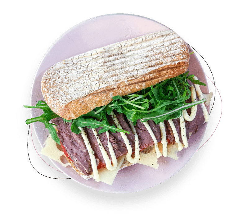Ein Sandwich auf einem rosa Teller, gefüllt mit Rindfleischscheiben, Rucola, Schweizer Käse und beträufelt mit Mayonnaise, serviert in einem rustikalen Ciabatta-Brötchen.