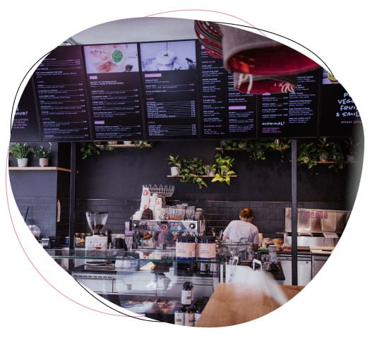 A barista works behind the counter of a modern café with digital menu boards above, surrounded by plants and coffee-making equipment.