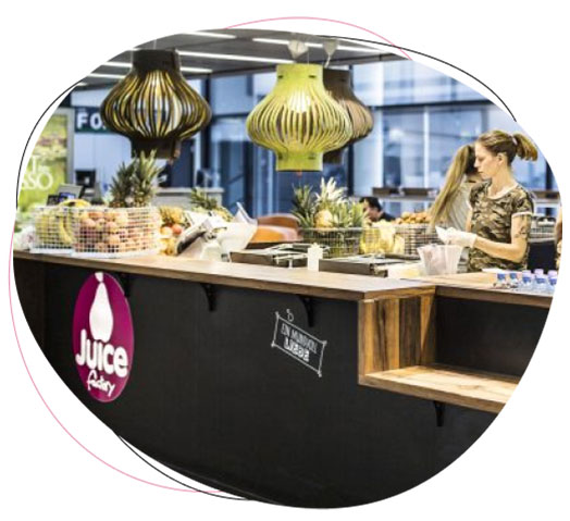 A woman stands behind a counter at a modern juice bar, preparing a drink. The counter displays fresh fruit, and stylish pendant lights hang overhead. The juice bar logo is visible on the front of the counter.