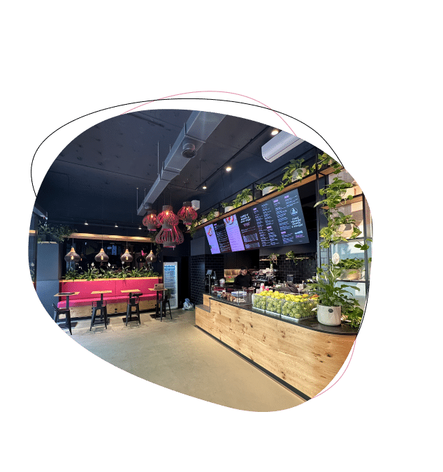 Modern café interior with wooden counter, digital menu boards, leafy plants, and a seating area with red booths and black stools under hanging red lamps. The space is well-lit and stylishly decorated.